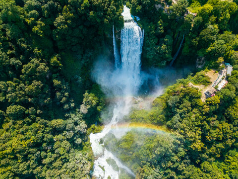 Aerial view of the cascading Marmore Falls plunges amidst lush green foliage, creating a vibrant rainbow, Terni, Umbria, Italy.