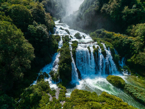 Aerial view of cascading waters plunge down the tiered rocks of Marmore Falls, embraced by verdant foliage, a scene of nature's raw power, Terni, Umbria, Italy.