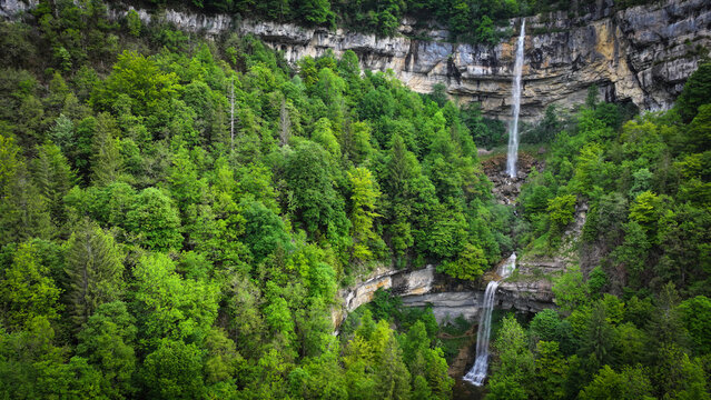 Aerial view of cascading Queue du Cheval waterfalls plunging down rocky cliffs amidst a vibrant tapestry of emerald and deep green forests, Saint-Claude, Bourgogne-Franche-Comte, France.