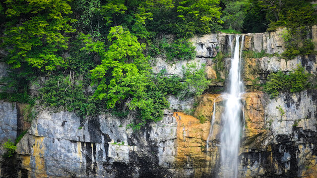 Aerial view of the cascading waterfall plunges from the cliff face, framed by vibrant green trees against the rugged, textured rock, Saint-Claude, Bourgogne-Franche-Comte, France.
