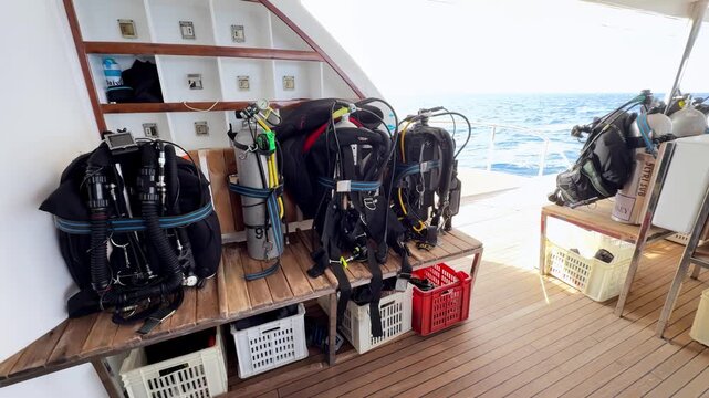 Professional scuba diving equipment including BCDs and air tanks organized on a wooden bench of a luxury liveaboard boat sailing across the bright blue Red Sea during a sunny summer morning expedition