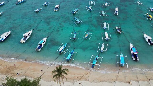 Overhead drone footage of traditional outrigger boats and speedboats anchored in clear turquoise tropical water beside a sandy beach lined with palm trees and gentle waves.