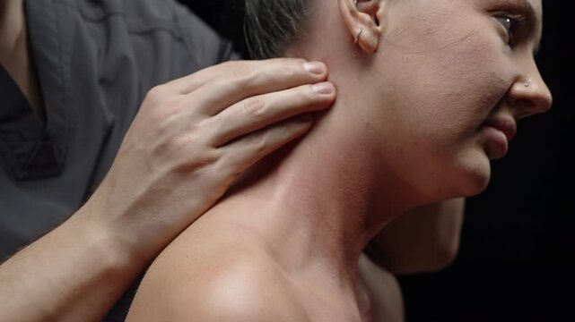 Close-up of professional hands performing therapeutic neck and shoulder massage on a seated person, easing tension with precise pressure techniques against a dark studio background