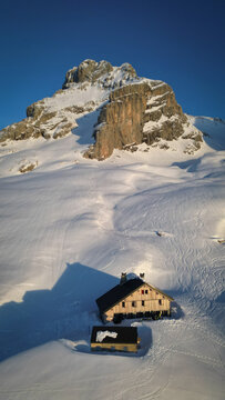 Aerial view of a Refuge de la Pointe Percee nestled in the crisp, snow-laden valley beneath the towering, rocky Pointe Percee, Le Grand-Bornand, Auvergne-Rhone-Alpes, France.