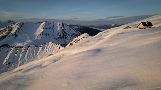 Aerial view of Refuge de la Pointe Percee in a snow-blanketed landscape under a soft sky, with mountain ranges in the distance near quaint huts, Le Grand-Bornand, Auvergne-Rhone-Alpes, France.