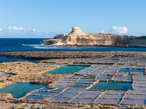 Aerial view of geometric salt pans reflecting the azure sky contrast with the rugged coastline and the distant, imposing rock formation, Zebbug, Malta.