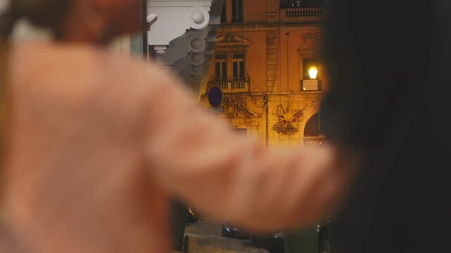 Over the shoulder shot of a blurry couple walking down a narrow, illuminated European street at night. Focus is on the atmospheric, historic architecture in the background