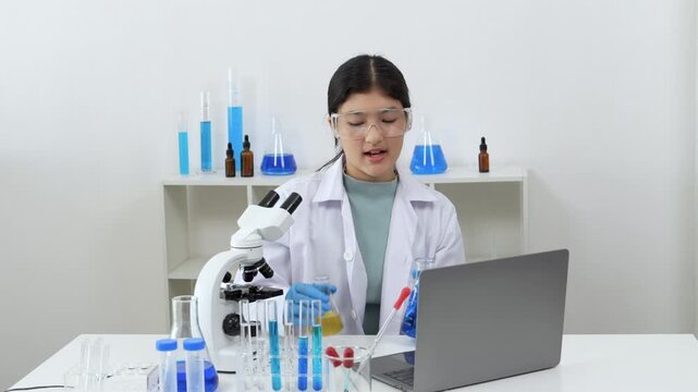 Young Asian women scientist working with test tube in laboratory. Scientist in a lab coat and safety glasses handles a test tube at a desk in a laboratory, make note on laptop, experimentation 