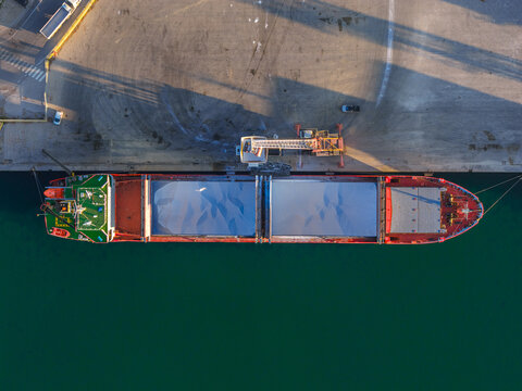 Aerial view of a cargo ship docked at the port, its red hull contrasting with the teal water and the pale concrete of the pier, Vasto, Abruzzo, Italy.