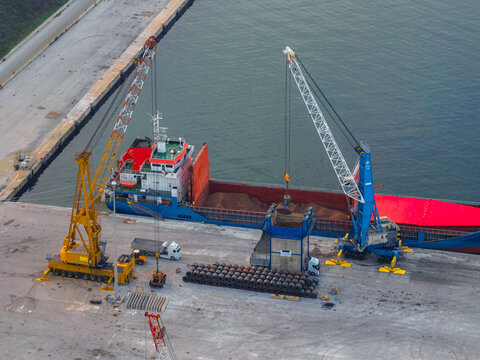 Aerial view of cranes towering over a cargo ship, its red deck contrasting with the blue hull, as it unloads its contents onto the dock, Vasto, Abruzzo, Italy.