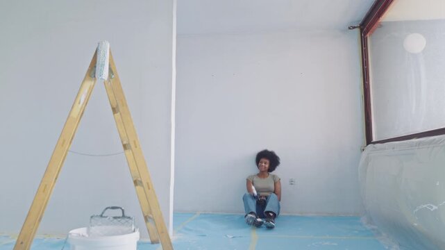 A young woman with an afro sits on a blue tarp in a sunlit room under renovation, surrounded by tools showing respite amidst painting and construction chaos.