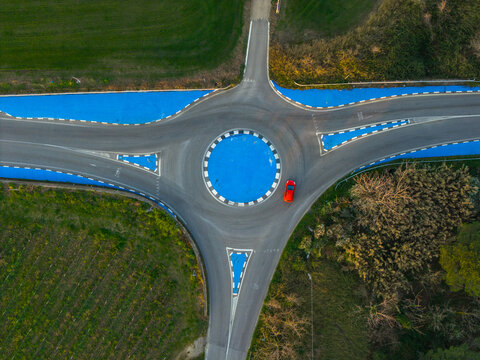 Aerial view of a vibrant red car navigating a blue and white roundabout, amidst lush greenery, creating a striking contrast, Vasto, Abruzzo, Italy.