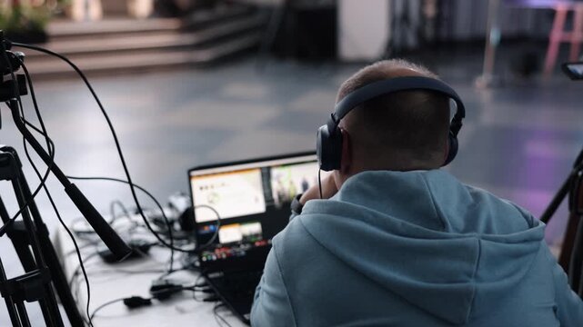 video streaming, technical director, camera rig. Man wearing headphones runs live video stream on laptop while monitoring camera setup at indoor professional conference.