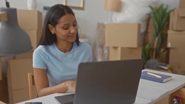 Young brunette woman typing on laptop with hands on keyboard amid stacked moving boxes in a building, smiling and leaning at desk; contentment fresh start.