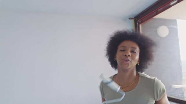 A woman with an afro hairstyle energetically paints a room, transforming its interior. She's focused, wearing casual attire and gloves, working on a wall near a window.