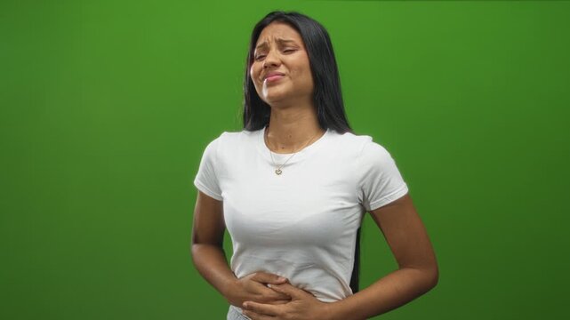 Young hispanic brunette woman clutching stomach, hands pressing abdomen, grimacing in studio green screen; pain nausea.