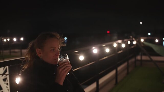A young woman in a dark jacket on a rooftop bar or terrace at night sips a glass of beer while looking pensively into the distance, surrounded by warm string lights and a relaxed ambiance, vintage