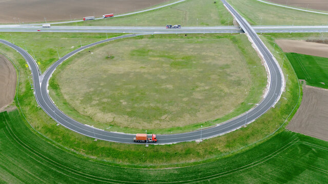 Aerial view of a looping highway interchange with trucks traversing the overpass and curved roads amidst green fields, Sremska Mitrovica, Vojvodina, Serbia.