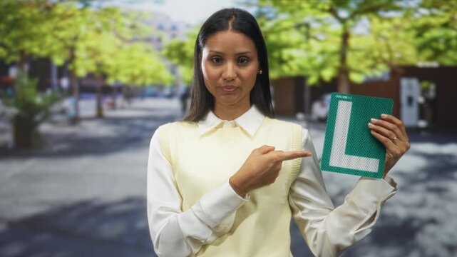 Woman pointing finger to green learner l sign while holding it at chest level on a sunlit street plaza with trees and buildings visible; confidence.