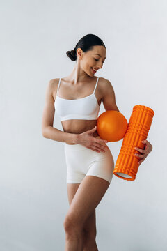 Slim female fitness model in white activewear holding an orange foam roller and ball, posing against a light gray background in a studio setting