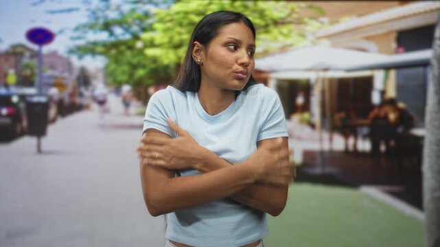Young hispanic woman hugging herself with crossed arms and closed eyes on a busy street cafe terrace; anxiety self comfort.