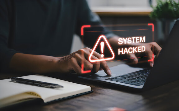 A person's hands interact with a laptop displaying a prominent red caution sign and the text message 'SYSTEM HACKED' in a dark office setting.
