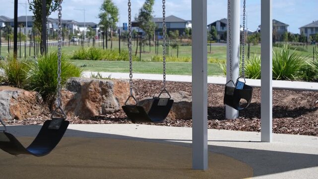 Shaded swing set in a playground in Manor Lakes, Australia, with modern suburban homes in the background. An empty and quiet, family-friendly outdoor space in Melborune's outer suburb.