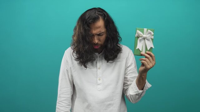 Man holding green gift box with white bow, frowning and looking down in teal studio; disappointment reflection.