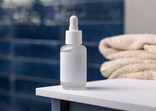 Dropper bottle with blank label near beige bath towels in blue bathroom, cosmetic mockup