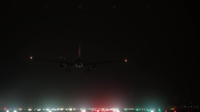 A commercial passenger airplane with bright navigation and landing lights descends onto a fully illuminated airport runway at night, completing its journey
