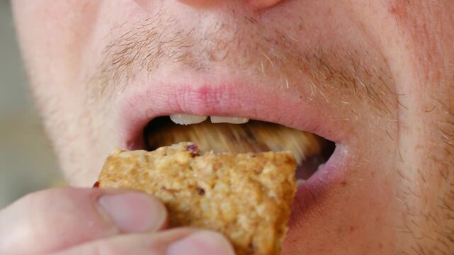 Extreme close up man mouth with visible herpes cold sore on his lip, bringing cereal bar to his mouth, taking bite, and chewing with his mouth closed. Close up of man with herpes eating cereal bar