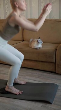 Sport fitness model recording squat workout at home using smartphone camera. Woman training on mat in living room, creating online tutorial, focusing on posture and strengthening lower body muscles
