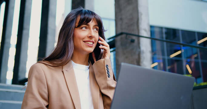 Businesswoman working on laptop at an outdoor while having a business call on phone. Focused woman having online meeting sitting on stairs near office building,