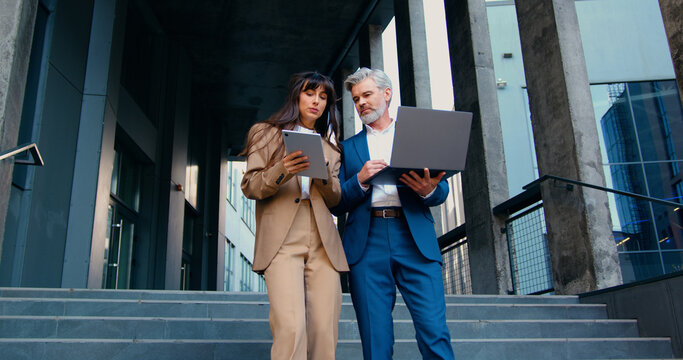 Two business professionals, a man and a woman in formal suits, walk down the stairs outside a modern office building after work, engaged in conversation. The woman uses a digital tablet, while the man