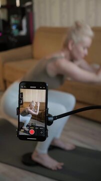 Female fitness trainer filming home workout on smartphone while doing squats on mat in living room. Sport woman coach creating tutorial content, demonstrating proper technique for legs, glute muscles