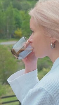 Female Savoring Aromatic Beverage On Balcony. Woman With Silver Earrings Tastes Coffee Overlooking Green Landscape. Lady In White Robe Enjoys Aromatic Crema From Glass Cup With Forest View