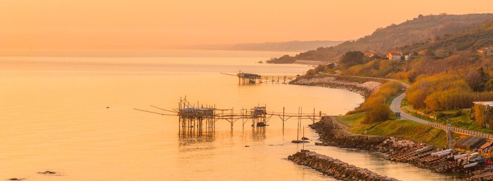 Aerial view of golden light bathing the Trabocchi coast, where ancient fishing platforms meet the winding coastal road, San Vito Chietino, Abruzzo, Italy.