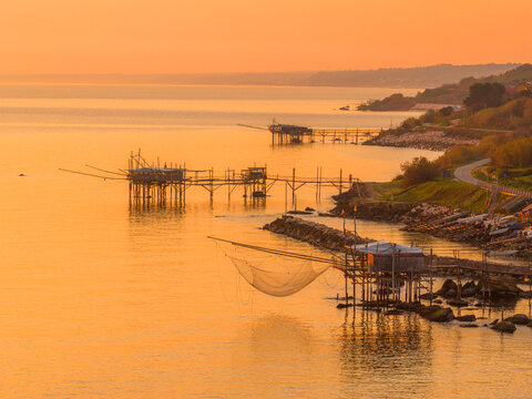 Aerial view of golden light bathing the Trabocchi along the rocky coastline, their nets shimmering above the tranquil sea, San Vito Chietino, Abruzzo, Italy.
