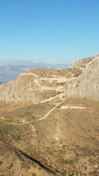 Aerial view of the ancient Acrocorinth fortress atop a rugged hill under a clear blue sky in Greece. The historic stone walls and pathways wind through the arid landscape.