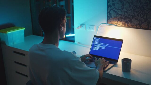 Programming at night as man types code on laptop at desk under blue light. Programming at night concept shows focused software development, late work, coding environment and modern technology workflow