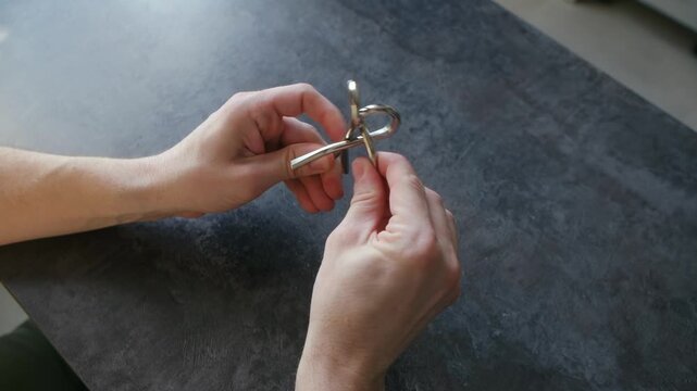Male hands manipulating shiny metal wire puzzle on dark tabletop, demonstrating problem solving, logic, concentration skills in an effort to find solution. Person solving metal brain teaser puzzle
