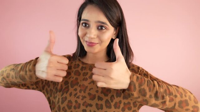 Close up of happy cheerful young Asian woman looking at camera showing thumbs up hands gesture like, ok, approve, agree, positive feedback, isolated against pink studio background.