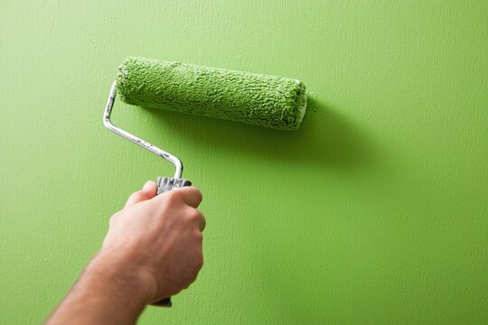 Hand holds a paint roller covered in bright green paint while rolling it across a smooth wall surface. Vivid green color fills the entire frame, suggesting an active home renovation in progress