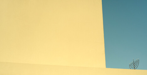 Obraz premium Minimalist composition of a geometric yellow wall edge against a blue clear sky, with a menorah silhouette on the roof.