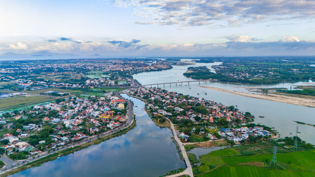 Aerial view of bridges elegantly cross the winding river, reflecting the sky's pale hues amidst the city's vibrant, compact buildings, Hoi An, Quang Nam, Vietnam.