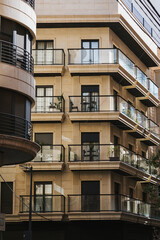 Obraz premium Contemporary city apartment building facade with multiple glass balconies, plants, and closed shutters, captured in warm afternoon sunlight on a quiet residential street corner.