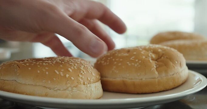 Close-up of a hand picking up a sesame hamburger bun from a plate in a kitchen.