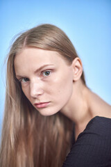 Fototapeta premium A portrait of a young woman with light brown hair and a neutral expression. She is wearing a black top on a soft blue background. Her gaze is direct, calm, and composed.