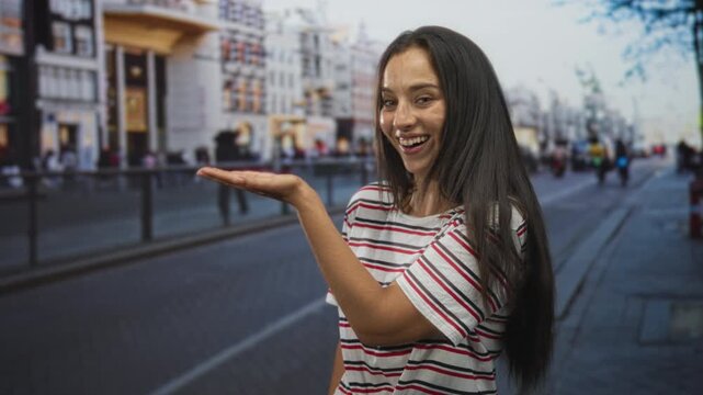 Young hispanic woman presenting palm up in striped shirt on busy city street while smiling and looking at hand; joy playfulness.