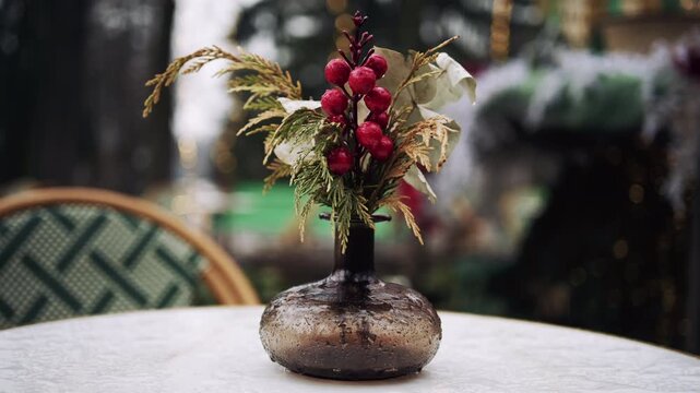 Small decorative glass vase with winter berries and greenery on outdoor table with festive background lights